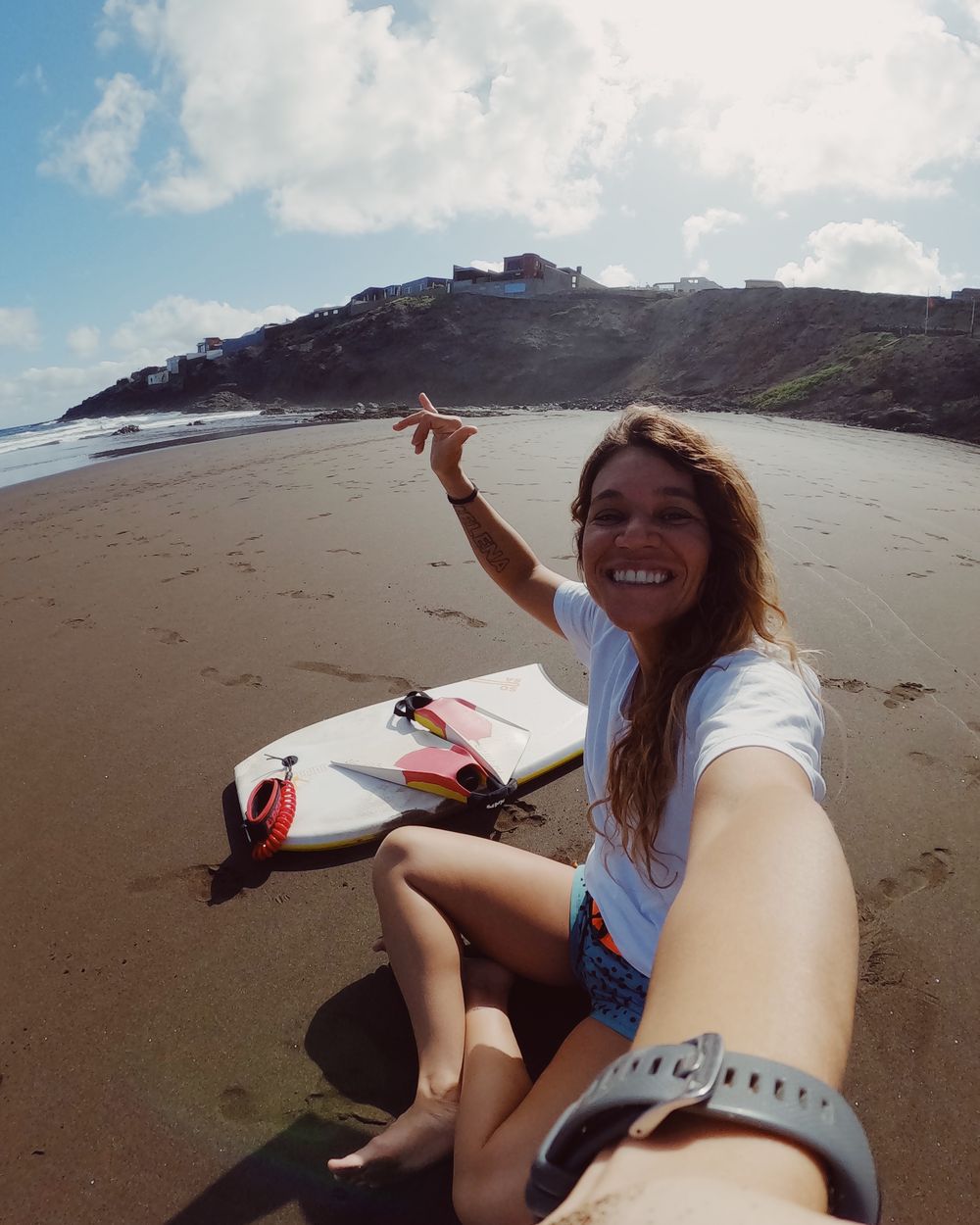 Isabela Sousa sentada na areia ao lado da prancha, sorrindo.
