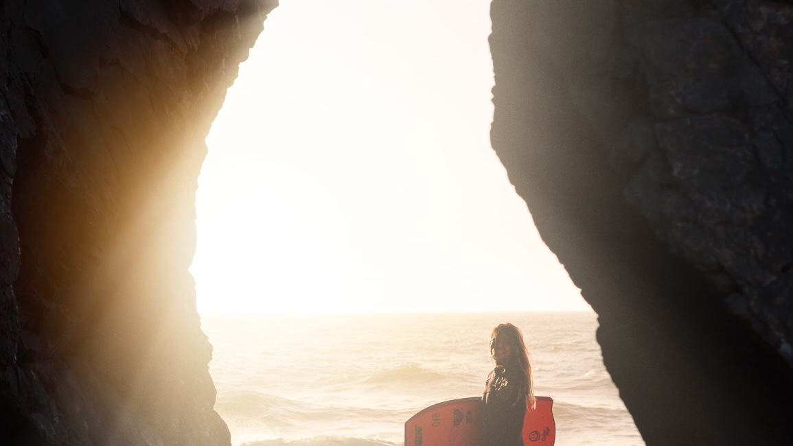 Silhueta de Isabela Sousa com a prancha dentro de um arco de pedra, ao pôr do sol.