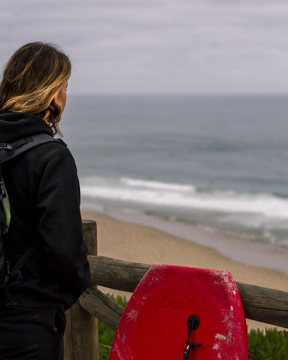 Isabela Sousa de costas, com mochila e pranchas, olhando para o mar.